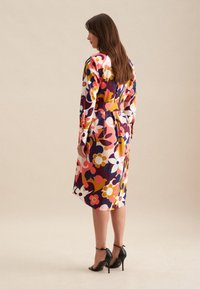 Kleid mit Blumenmuster, tailliert, lange Ärmel und knielanger Saum. Die Farben umfassen Rosa, Lila, Marineblau und Gelb. Schwarze Absätze.