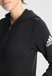 Haut à capuche zippé noir avec fermeture éclair à l'avant, poignets côtelés et logo Adidas blanc sur la manche gauche. Tissu lisse avec une coupe décontractée.