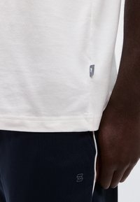 Κοντινό πλάνο ενός σκουρόχρωμου μπράτσου δίπλα σε λευκό πουκάμισο και σκούρο μπλε παντελόνι με μικρό λογότυπο και λευκή πλαϊνή ρίγα.