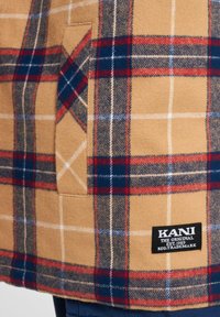 Plaid wool blend coat in tan, featuring red and blue check patterns, a patch pocket, and a black brand label stitched at the hem.