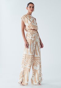 Une combinaison beige et blanche présentant un design à jambes larges, une taille élastique et un motif abstrait. Le haut a une coupe asymétrique et des manches ballons.
