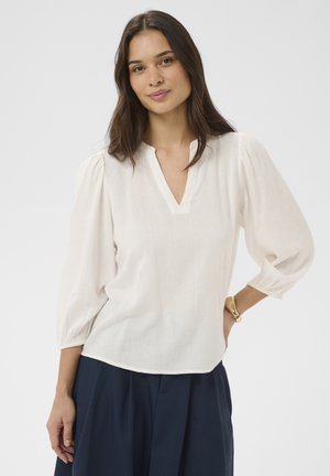 Kobieta o długich ciemnych włosach, ubrana w białą bluzkę z bufkami na rękawach i granatowe spodnie, stoi z jedną ręką na biodrze.