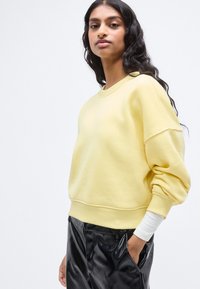 Κίτρινο βαμβακερό φούτερ με χαλαρή εφαρμογή, φαρδιά μανίκια και ριμπ μανσέτες, συνδυασμένο με λαμπερό μαύρο παντελόνι. Λεία υφή, μινιμαλιστικός σχεδιασμός.