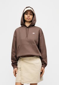 Καφέ φούτερ με κουκούλα και μπροστινή τσέπη, με λευκό λογότυπο. Συνδυασμένο με ανοιχτό μπεζ τζιν φούστα με πλευρικό σκίσιμο.