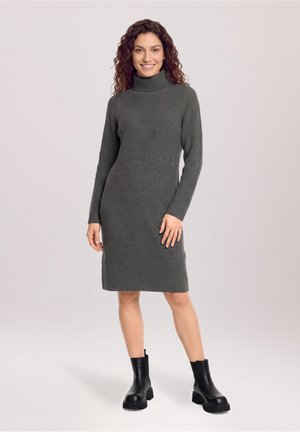 Femme debout et souriante portant une robe pull col roulé côtelée grise et des bottines noires sur un fond clair uni.