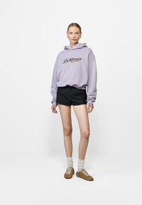 Lila kapucnis pulóver, rajta "La Mania" felirattal, fekete csíkos rövidnadrág, fehér zokni és barna velúr cipő, egy sima háttér előtt áll.