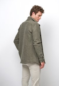 Olivgrön ylleblandad jacka med krage, knäppning framtill och sidofickor. Har långa ärmar och en något rundad fåll.