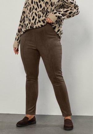 Braune Faux-Suede-Hosen mit schlanker Passform, ausgestattet mit Vordernähten und einer glatten Textur, kombiniert mit dunkelbraunen Slippern.