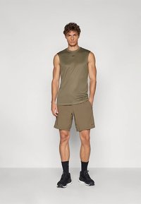 Oliwkowo-zielony sportowy top bez rękawów w zestawie z dopasowanymi szortami, oba wykonane z lekkiego materiału. Czarne sneakersy dopełniają strój.