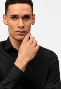 Chemise noire à manches longues avec un design ajusté, un léger éclat et des poignets à boutonnage. Le modèle touche son menton, mettant en valeur sa peau lisse et ses cheveux courts.
