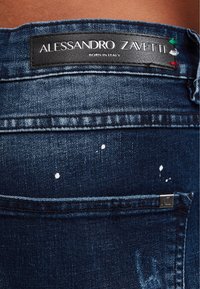 Mörkblå denimjeans med en svart läderetikett som står "Alessandro Zavetti." Har vita färgfläckar och silverfärgade detaljer.