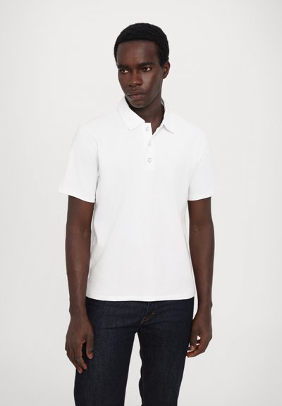 Polo shirt blanc à manches courtes avec patte de boutonnage à trois boutons, fabriqué en coton doux. Porté avec un jean en denim bleu foncé.