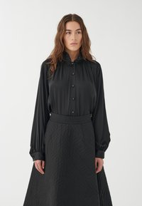 Blusa negra con cierre de botones y cuello alto, combinada con una falda larga negra acolchada que presenta un patrón geométrico. Textura suave.