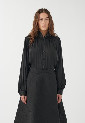Schwarze Bluse mit Knopfverschluss und hohem Kragen, kombiniert mit einem langen schwarzen gesteppten Rock mit geometrischem Muster. Glatte Textur.