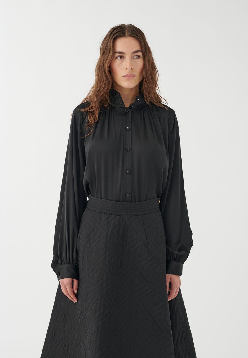 Blusa negra con cierre de botones y cuello alto, combinada con una falda larga negra acolchada que presenta un patrón geométrico. Textura suave.