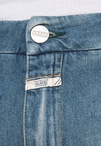 Blå denimjeans med en silverknapp med "CLOSED" ingraverat, och en vit tygetikett sydd inuti midjebandet.