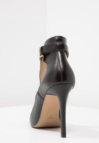 Černá kožená lodička na vysokém podpatku s páskem kolem kotníku, špičatou špičkou a lesklým povrchem. Má úzký jehlový podpatek a hladký vnitřek.