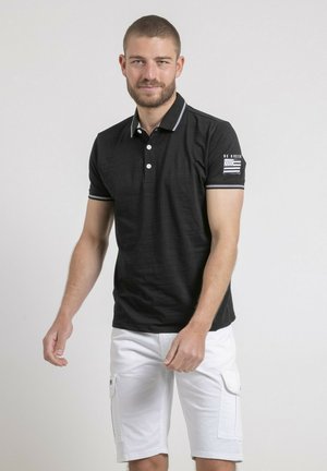 Homme aux cheveux courts et barbe, portant un polo noir avec un emblème sur la manche et un short cargo blanc, debout devant un fond uni.