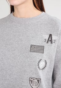 Grijze sweatshirt met verschillende geborduurde patches: een Amerikaanse vlag, gevleugelde letters "A2," een lauriertak-symbool en een adelaar-embleem.