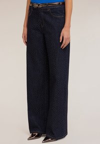 Kvinna som bär vida mörk denimjeans med små rhinsten detaljer, en blå topp, bälte och spetsiga metalliska högklackade skor.