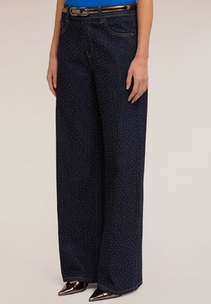 Femme portant un jean large en denim foncé avec de petits détails en strass, un haut bleu, une ceinture et des chaussures à talons aiguilles métalliques.