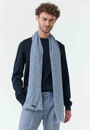 Mann mit lockigem Haar, der einen marineblauen Pullover, einen hellblauen gemusterten Schal und hellgraue Hosen trägt, steht mit den Händen in den Taschen.