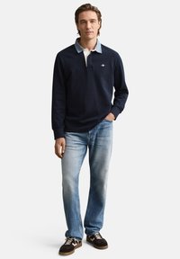 Marineblauw poloshirt met lange mouwen en een lichtblauwe kraag, gecombineerd met verweerde blauwe spijkerbroek en donkere sneakers. Opvallende borduursels op de borst.