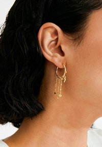 Kuldne rõngaskõrvarõngas ripatsitega ahelatega, millel on väikesed võlusümbolid, sealhulgas süda ja täht, eksponeeritud tekstureeritud naha taustal.