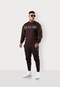 Dunkelbrauner Sweatshirt und passende Jogginghose mit dem weißen "ESTEEM"-Logo. Weiße Sneaker, runde Sonnenbrille, sichtbare Tattoos an den Armen.