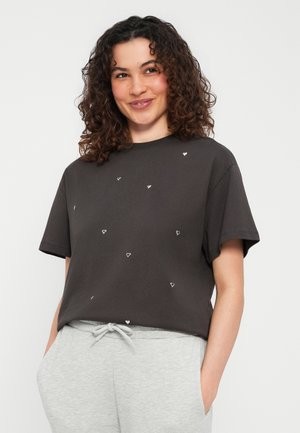 Femme aux cheveux bouclés portant un t-shirt noir à petits motifs de cœurs blancs et un pantalon gris clair à cordon, souriante et posant.