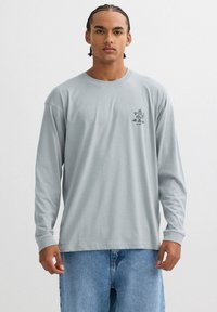 Camiseta de manga larga gris claro hecha de algodón con cuello redondo, que presenta un pequeño gráfico negro en la parte izquierda del pecho.