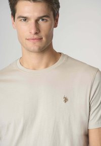 T-shirt beige in cotone con collo rotondo e maniche corte. Presenta un piccolo logo ricamato sul lato sinistro del petto. Tessuto morbido.