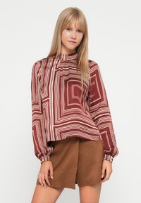 Bordeauxfarbige Bluse mit weißen geometrischen Mustern, hohem Kragen und langen Ärmeln, kombiniert mit braunen Wildledershorts mit elastischem Saum.