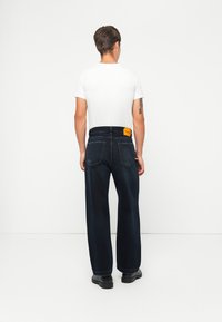 Dark blue wide-leg jeans with a light tan tag on the waistband. The model wears a plain white t-shirt and black shoes, viewed from the back.