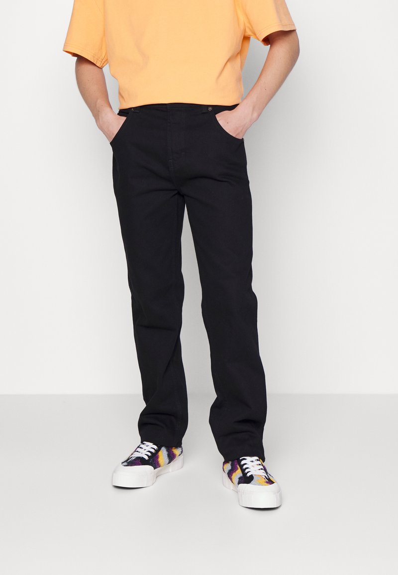 Pantalones negros de pierna recta combinados con una camiseta corta naranja y zapatillas multicolores con un diseño estampado, que presentan un clásico diseño de cinco bolsillos.