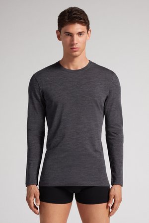 Jeune homme portant une chemise moulante gris foncé à manches longues et un short noir, debout devant un fond clair uni.