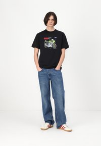 Zwarte t-shirt met motorfietsafbeelding, ronde hals, korte mouwen; gecombineerd met losse blauwe jeans en witte sneakers met rode strepen.