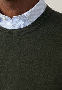 Maglione in maglia verde scuro con collo a coste, indossato sopra una camicia azzurra chiara con un motivo geometrico.