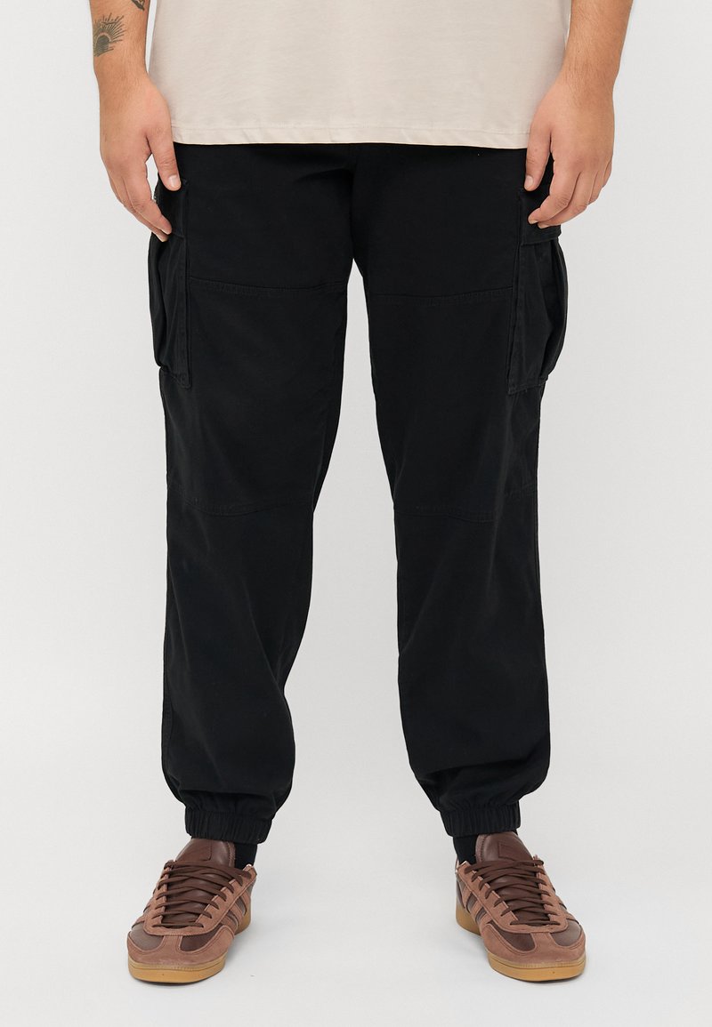 Zwarte cargobroek met een ontspannen pasvorm, elastische manchetten en zijzakken. Gemaakt van een gladde stof met een matte afwerking. Gecombineerd met bruine sportschoenen.