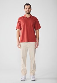 Röd kortärmad polo t-shirt med krage och en framficka, ihop med beiga byxor och vita sneakers på en neutral bakgrund.