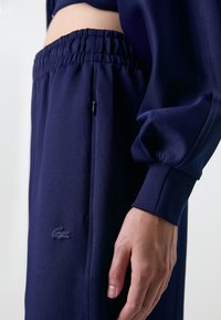 Calças de desporto azul-marinho com um cós elástico, bolso lateral e tecido texturizado. Apresenta um pequeno logótipo perto da bainha.