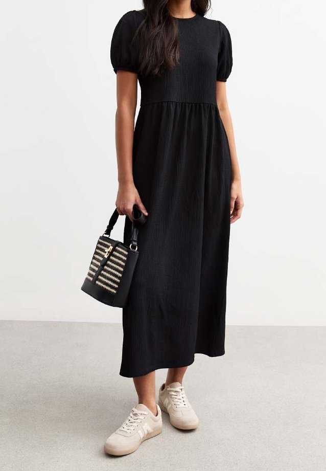 Femme portant une robe midi noire à manches courtes bouffantes, des baskets blanches et tenant un petit sac à main noir et beige à motifs.