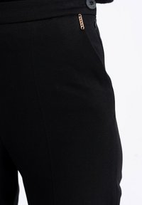 Zbliżenie czarnych spodni pokazujących boczną kieszeń z małym złotym łańcuszkiem i czarnym guzikiem w talii.