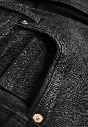 Primo piano di una tasca di jeans in denim nero con cuciture visibili e due piccoli rivetti in ottone vicino al bordo della tasca.
