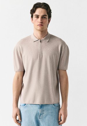 Polo de manga corta en beige claro, con textura acanalada, con cierre delantero y cuello plano. Combinado con pantalones cortos de mezclilla azul claro.