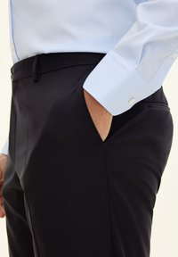Schwarze Hosen mit glatter Textur, ausgestattet mit einer Seitentasche. Kombiniert mit einem hellblauen Hemd mit Knöpfen, das Knopfmanschetten zeigt.