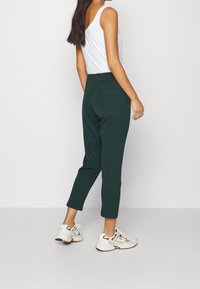 Dunkelgrüne Hosen mit taillierter Passform, elastischem Bund und Seitentaschen, kombiniert mit einem weißen Tanktop und beigen Turnschuhen.