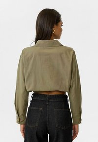 Afgekorte, olijfgroene shirt met een kraag en lange mouwen, gecombineerd met hooggesneden donkere spijkerbroeken met achterzakken.