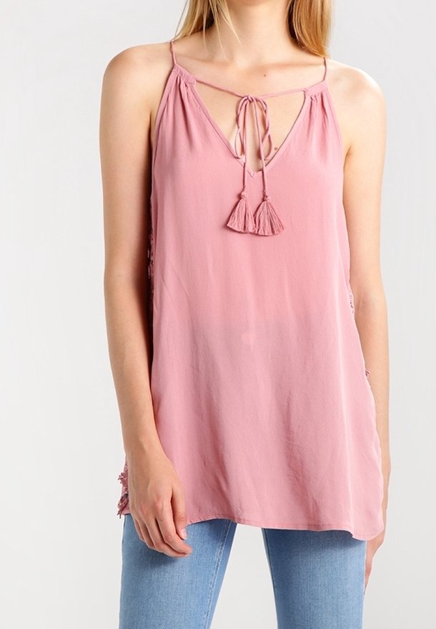 Femme portant un haut rose sans manches avec des bretelles fines et des liens à pompons au niveau de l'encolure, associé à un jean bleu clair.