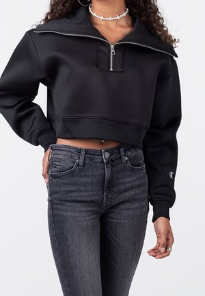 Kvinde iført en sort cropped sweatshirt med kvart lynlås og mørkegrå high-waisted jeans, accessoriseret med en perle choker og hoop øreringe.